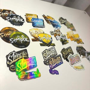 Puede incluir: Una colección de pegatinas coloridas con la palabra "Simple" en varios diseños. Algunas pegatinas presentan texto adicional, incluyendo "MLG Columbus 2016", "Champion PGL Stockholm 2021" y "ESL". Las pegatinas tienen un acabado brillante.