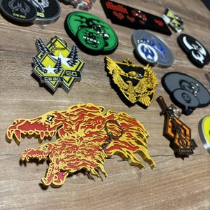 Puede incluir: Una colección de llaveros y pines coloridos con varios diseños. Los llaveros y pines presentan logotipos, emblemas y texto, incluyendo "CS GO" y "HYDRA". Los artículos son en tonos de amarillo, naranja, negro y verde.