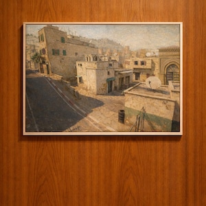 Puede incluir: Una impresión enmarcada de un detallado paisaje urbano, con edificios de paredes beige y contraventanas verdes. La obra de arte representa una escena callejera con una carretera y sombras, sobre un fondo de colinas. El marco es blanco y el fondo es una superficie de madera.