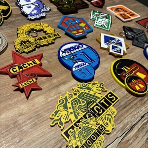 Puede incluir: Una colección de llaveros coloridos con varios diseños y texto, incluyendo una estrella roja con "CACHE" y un óvalo azul con "VERTIGO 51". Los llaveros están sobre una superficie de madera.