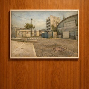 May include: A framed print depicting a detailed, realistic scene of an urban environment. The artwork showcases a gritty, industrial area with buildings, containers, and a paved road. The color palette is muted, with shades of gray, brown, and blue.