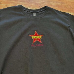 以下が含まれることがあります： 赤と黄色の星のデザインが施された濃いグレーのTシャツ。「CACHE」の文字が黄色の長方形に、そして「de_cache」の文字が星の下にピンク色で書かれています。柔らかい素材で作られたTシャツです。