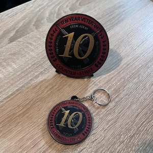 Könnte beinhalten: Eine runde Ten Year Veteran Counter-Strike Challenge Coin und ein passender Schlüsselanhänger. Beide Artikel haben eine schwarze Mitte mit einer großen goldenen 10, gekreuzten Gewehren und roten Rändern mit dem Text Ten Year Veteran, Dedication, Community, Teamwork, Tactics.