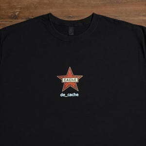 Könnte beinhalten: Schwarzes T-Shirt mit einem roten Stern-Grafik. Der Stern hat das Wort "CACHE" in einem rechteckigen Feld. Unter dem Stern steht der Text "de_cache" in Hellblau. Das T-Shirt ist aus weichem Material.