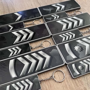 May include: A collection of rectangular keychains featuring a dark grey camouflage pattern. Each keychain displays silver-coloured chevron rank insignia, with some designs including a silver star emblem. Each piece is attached to a metal ring.
