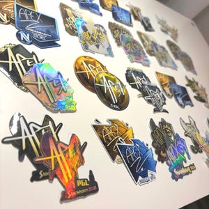 Puede incluir: Una colección de pegatinas APEX con varios diseños y colores, incluyendo elementos dorados, azules y holográficos. Algunas pegatinas presentan texto como "DreamHack 2016" y "PGL Stockholm 2021".