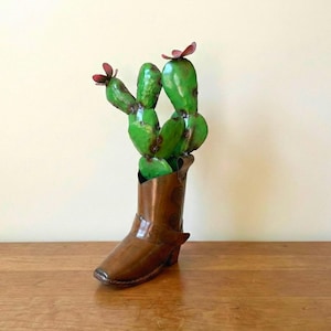 May include: A metal cactus sculpture with green leaves and red flowers, displayed in a brown cowboy boot. The boot has decorative details and a small heel, creating a rustic, southwestern-themed decorative piece.