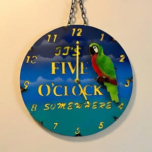 May include: Round wall clock with a blue and green gradient background. The clock face features the text "JJ's FIVE O'CLOCK SOMEWHERE" in yellow. A green, red, and black parrot is perched on the right side. The clock is suspended by a black chain.