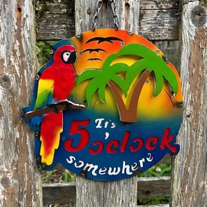 May include: A round metal sign with a parrot, palm tree, and the text "It's 5 o'clock somewhere." The sign features a colorful sunset design with a blue ocean and birds in flight. It hangs from a chain.