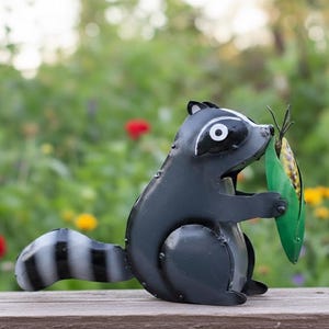 May include: A metal raccoon figurine in shades of gray and black, holding a green corn husk with a yellow and brown corn cob. The raccoon has a striped tail and is set against a blurred background of greenery and flowers.
