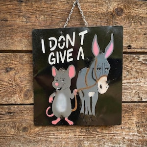 May include: Black metal sign with the phrase "I DON'T GIVE A" in white, featuring a cartoon mouse holding a donkey's lead. The mouse is gray with pink ears and feet. The donkey is gray with a white muzzle and pink ears.