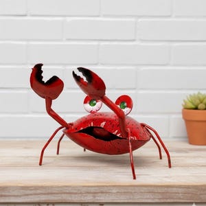 May include: A red metal crab sculpture with large claws and googly eyes. The crab has a textured finish and is posed on a wooden surface. The crab's eyes are white with green centers. A decorative item for coastal or nautical themes.