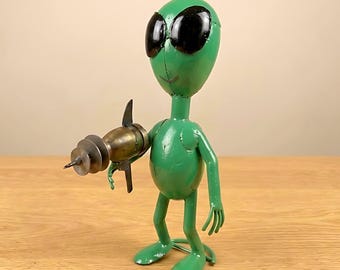 Op de afbeelding: Een groene metalen alien sculptuur met grote zwarte ogen en een straalwapen. De alien houdt een bronskleurig straalwapen vast. De sculptuur staat op een houten oppervlak.