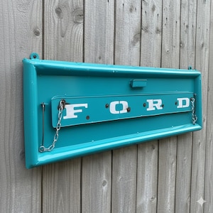 Op de afbeelding: Een turquoise metalen bord met het woord "FORD" in witte blokletters. Het bord heeft een rechthoekige vorm met een gebogen boven- en onderkant. Het is gemonteerd op een houten achtergrond.