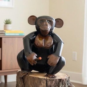 May include: A handcrafted, black and brown chimpanzee sculpture, seated on a tree stump. The sculpture has a glossy finish and holds a small, orange-tipped object. The chimpanzee's face and chest are a lighter brown color. The sculpture is a unique decorative item.