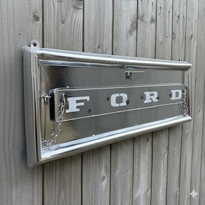 Op de afbeelding: Een glanzend metalen bord met het woord "FORD" in witte letters. Het rechthoekige bord heeft een zilveren afwerking en is bevestigd aan een houten hek. Kettingen hangen aan de zijkanten.