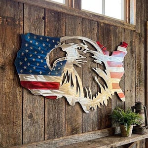 May include: Metal wall art depicting the United States map with an eagle silhouette. The flag design features red and white stripes and blue with white stars. The artwork is mounted on a rustic wooden wall.