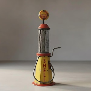 May include: A vintage-style gas pump replica in yellow, red, and silver. The pump features the Shell logo on top and the word "SHELL" vertically down the front. It has a handle and a mesh cylinder in the center, evoking a retro aesthetic.