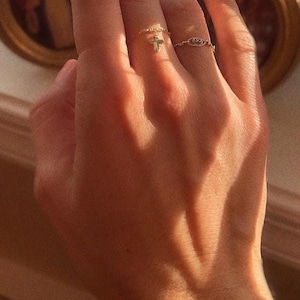 May include: Two gold-toned rings worn on the fingers. One ring features a small dangling cross charm attached to a delicate chain band. The second ring is a thin, open-work band with an oval-shaped decorative element in the centre.