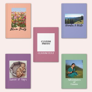 May include: Five notebooks with different covers. One has a field of flowers and the text "Bloom Daily". Another has a mountain scene and the text "Wander & Write". A third has a cat in a bed and the text "Queen of Naps". The fourth has a woman reading and the text "Adventures of Emma". The fifth has space for a custom photo and text.