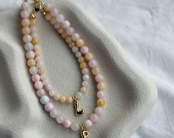 May include: Two layered beaded necklaces featuring smooth, round, pastel-coloured stones in shades of soft pink, peach, and cream. Each necklace includes a small, polished gold-tone metal letter charm, specifically the letters L and P, attached.