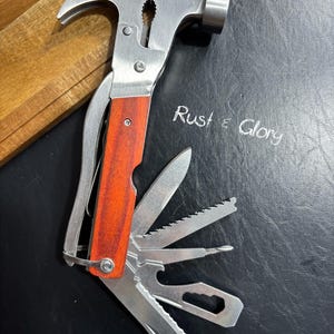 May include: A multi-tool hammer with a stainless steel head and a wooden handle. The tool includes a hammer, knife, saw, and other implements. The text "Rust & Glory" is written on the surface.