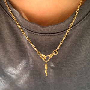 May include: A gold chain necklace featuring a small, curved horn-shaped pendant suspended from a swivel clasp. The necklace is constructed with elongated oval links and has a polished metallic finish, resting against a dark grey fabric background.