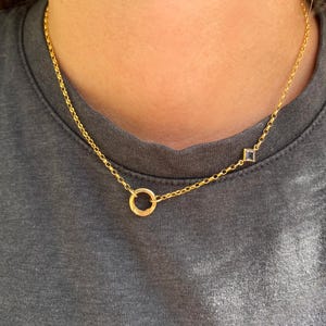 Op de afbeelding: Een goudkleurige ketting met een cirkelvormige ringhanger en een enkele kleine, vierkant geslepen, heldere edelsteen in een gouden zetting. De fijne schakelketting rust tegen de hals en benadrukt de metalen afwerking en de twee unieke bedels.