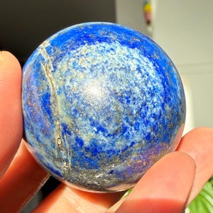 May include: A lapis lazuli sphere, held in a hand. The gemstone is a deep blue colour with lighter blue and gold flecks. The sphere is smooth and round, reflecting light. A decorative item.