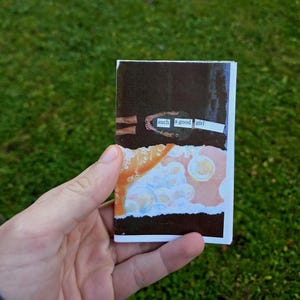 May include: A small, handmade collage with the text "such a good girl" on a white strip. The collage features torn paper with orange, brown, and white abstract designs. The background is green.