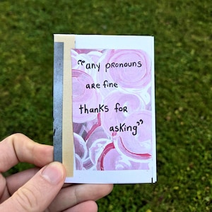 May include: A small, rectangular card with the text "any pronouns are fine thanks for asking" in black script. The background features a pink and white abstract painted design. The card is held against a backdrop of green grass.