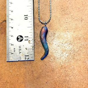 May include: A pendant necklace featuring a curved horn-shaped charm. The charm is approximately 1.5 inches long and displays a color gradient of blue, green, and copper. The necklace chain is a silver-colored ball chain.
