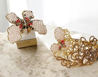 May include: Two handmade, ornate jewelry pieces. One is a ring with a floral design, featuring pink beads and gold wire. The other is a matching bracelet with a similar design. Both pieces have red and black accents.