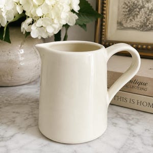 May include: A cream-colored ceramic pitcher with a smooth, glossy finish, a curved handle, and a small pouring spout. The pitcher has a simple, cylindrical body with a slightly wider base, presenting a clean and solid appearance for serving.