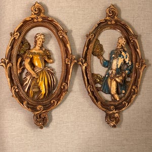 May include: Two vintage wall plaques featuring oval frames with ornate detailing. Each frame contains a relief sculpture of a person in period clothing. One figure wears a yellow dress, the other a blue coat. The frames are brown.