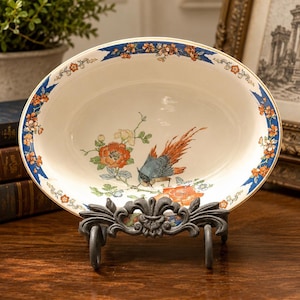 May include: An oval ceramic bowl featuring a cream-colored base with a central illustration of a bird with orange and blue plumage perched on a branch among orange flowers. The rim is decorated with a blue and orange floral pattern.