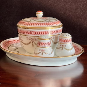 May include: A white porcelain serving set with red and gold accents. The set includes a lidded bowl, two shakers, and an oval tray. The pieces are decorated with a repeating red pattern and gold trim, creating an elegant aesthetic.