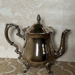 May include: A silver-colored teapot with a decorative handle and spout. The teapot has a lid with a pinecone-shaped finial and sits on three small feet. The polished surface reflects the surrounding environment.