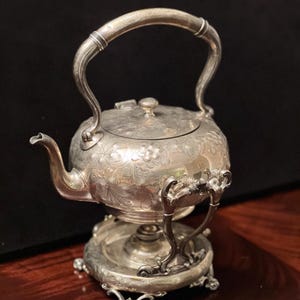 May include: A silver tea kettle with a floral design, a curved handle, and a matching stand. The kettle has a spout and a lid with a small knob. The stand has ornate legs and a circular base. The kettle is likely made of silver.