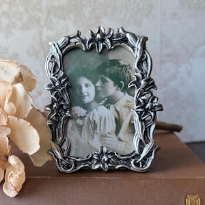 May include: Ornate silver-toned picture frame with a vintage photograph of two children. The frame features a floral design around the edges. The photograph is in sepia tones, adding to the antique aesthetic. A decorative piece for displaying cherished memories.