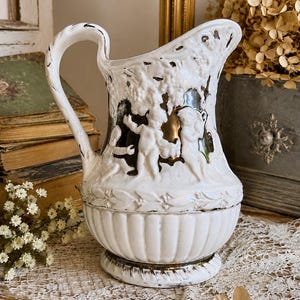May include: A white ceramic pitcher featuring a relief design of cherubs amidst foliage. The piece has a glossy finish with metallic gold accents on the figures and base. It includes a curved handle, a fluted lower body, and a decorative rim.