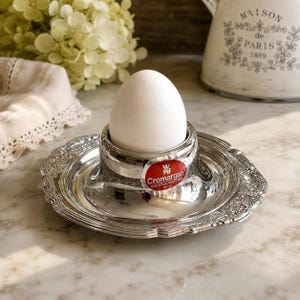 May include: A silver-colored metal egg cup with a wide, ornate, scalloped base and a smooth central holder. A red oval sticker on the front features the white text WMF Cromargan 18/10. A single white egg is positioned upright inside the cup holder.