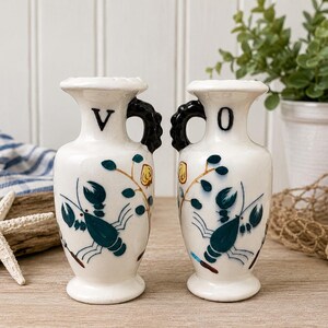 May include: Two white ceramic amphora-style vases with black handles. Each vase features a hand-painted teal lobster motif and a single black letter, V on the left and O on the right, positioned near the top rim. The vases have a flared neck.
