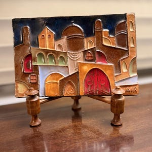 May include: A rectangular, colorful ceramic tile depicting a Middle Eastern cityscape, mounted on a wooden stand. The tile features buildings in shades of brown, orange, and red, with a blue sky background. The stand has three wooden legs.