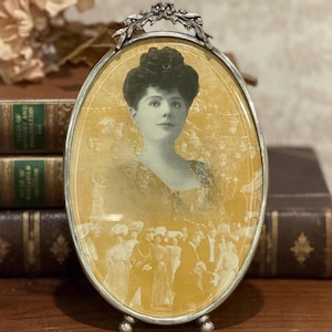 May include: Oval antique silver-toned picture frame with a portrait of a woman. The frame has a decorative floral design at the top and a stand at the base. The portrait is in sepia tones, with a background of a crowd of people.