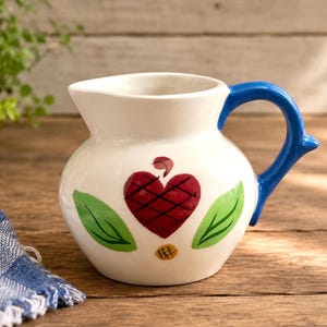 May include: A white ceramic pitcher featuring a hand-painted red heart with a cross-hatched pattern, flanked by two green leaves and a small yellow dot at the base. The pitcher has a glossy finish and a solid blue handle with a curved shape.