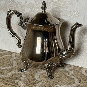 May include: A silver-colored teapot with ornate detailing, including a curved handle and a decorative spout. The teapot has a lid with a small, textured finial. It sits on three small feet and reflects the surrounding environment.