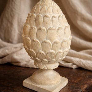 May include: A cream-colored decorative artichoke sculpture. The sculpture has a detailed, textured surface and is mounted on a square base. The artichoke design is a classic element for home decor.