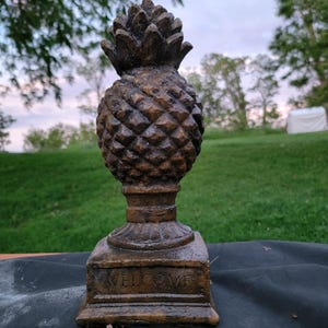 May include: A bronze-colored pineapple statue with a textured surface. The statue sits on a square base with the word "WELCOME" inscribed. The pineapple has a detailed crown and a classic design.