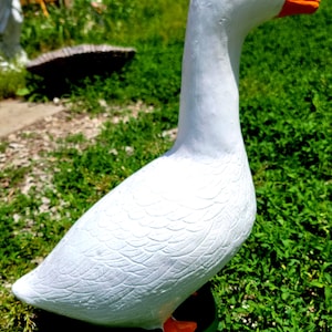 May include: A white goose figurine with a bright orange beak and orange feet. The sculpture features molded feather textures across its body and a smooth, rounded head with a small black eye. It stands on a dark green base, capturing the bird in a profile view with its neck extended upward.